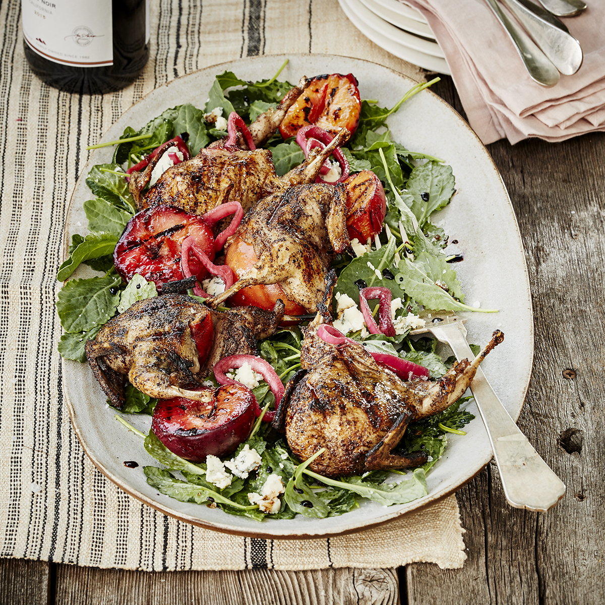 A close up of an entree with a bed of baby kale topped with roasted quail, plums, and Hibiscus Pickled Shallots.